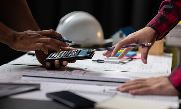 two hands using a calculator on a construction plan with a hard hat background discussing budget for four projects